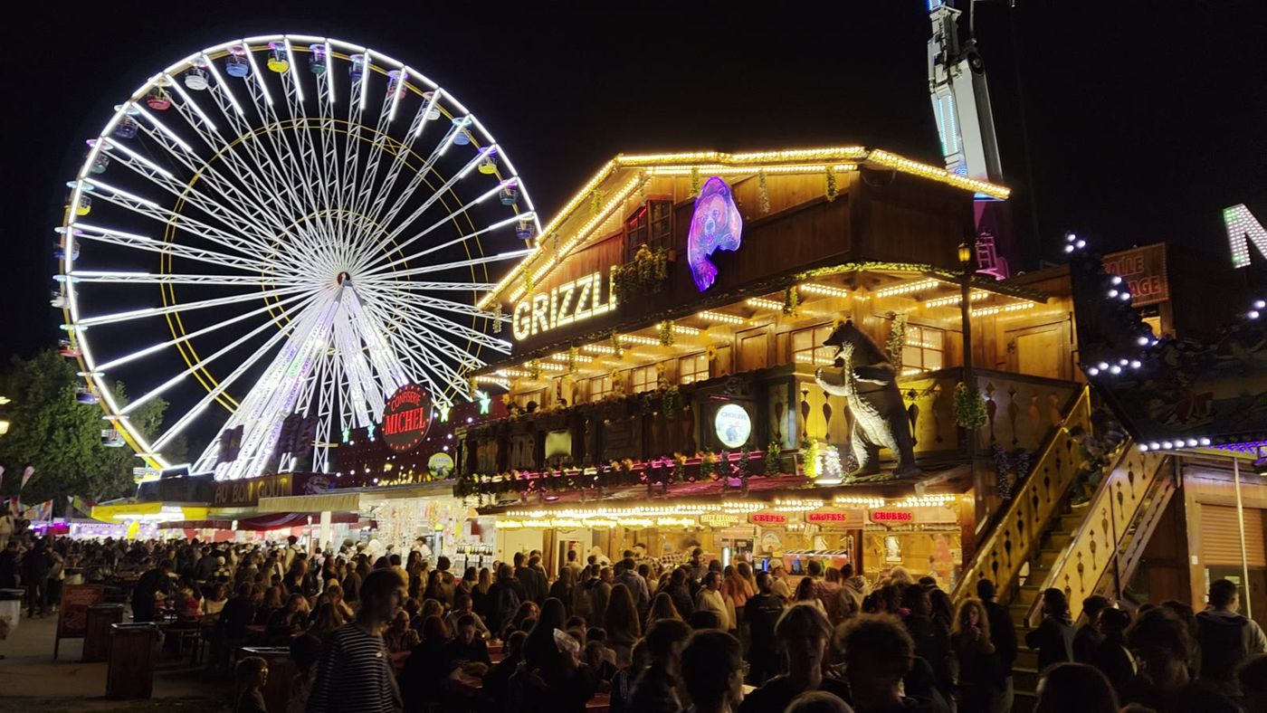 Foule dense devant la grande roue et un stand illuminé, de nuit